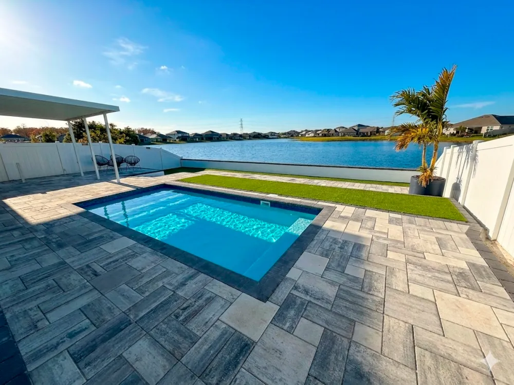 Deck de pavers cinza e brancos em volta de uma piscina retangular de frente para um lago, com grama sintética e uma palmeira