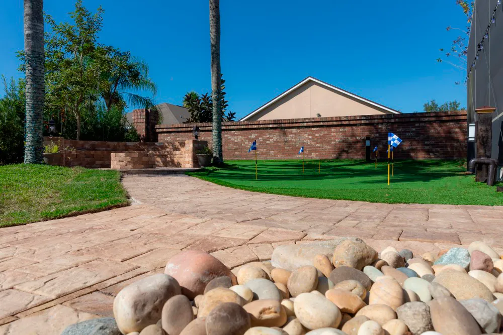 Patio de pavers color arena con piedras de río, un putting green y un muro de ladrillo en el jardín